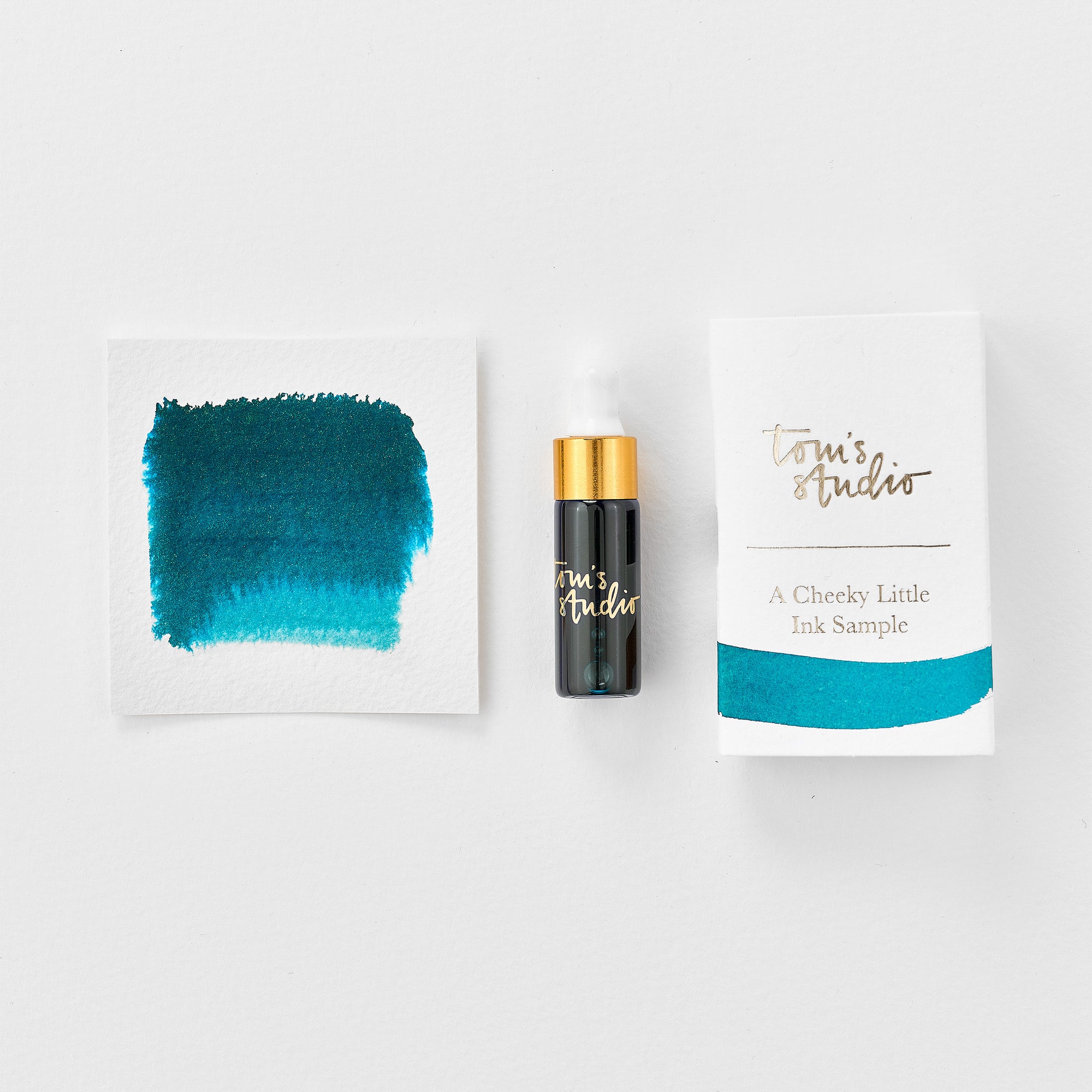 A mini ink sample bottle of Neptune blue fountain pen ink by Tom’s Studio, displayed alongside a swatch of the ink on textured paper and its matching packaging. The bottle is small, with a gold cap and a white pipette dropper, and features 'Tom’s Studio' in gold lettering. The ink swatch shows a rich, shimmering hue with a gradient from deep to lighter tones. The packaging, a small white card, has 'Tom’s Studio' embossed in gold at the top, with a brushstroke of the ink colour at the bottom.