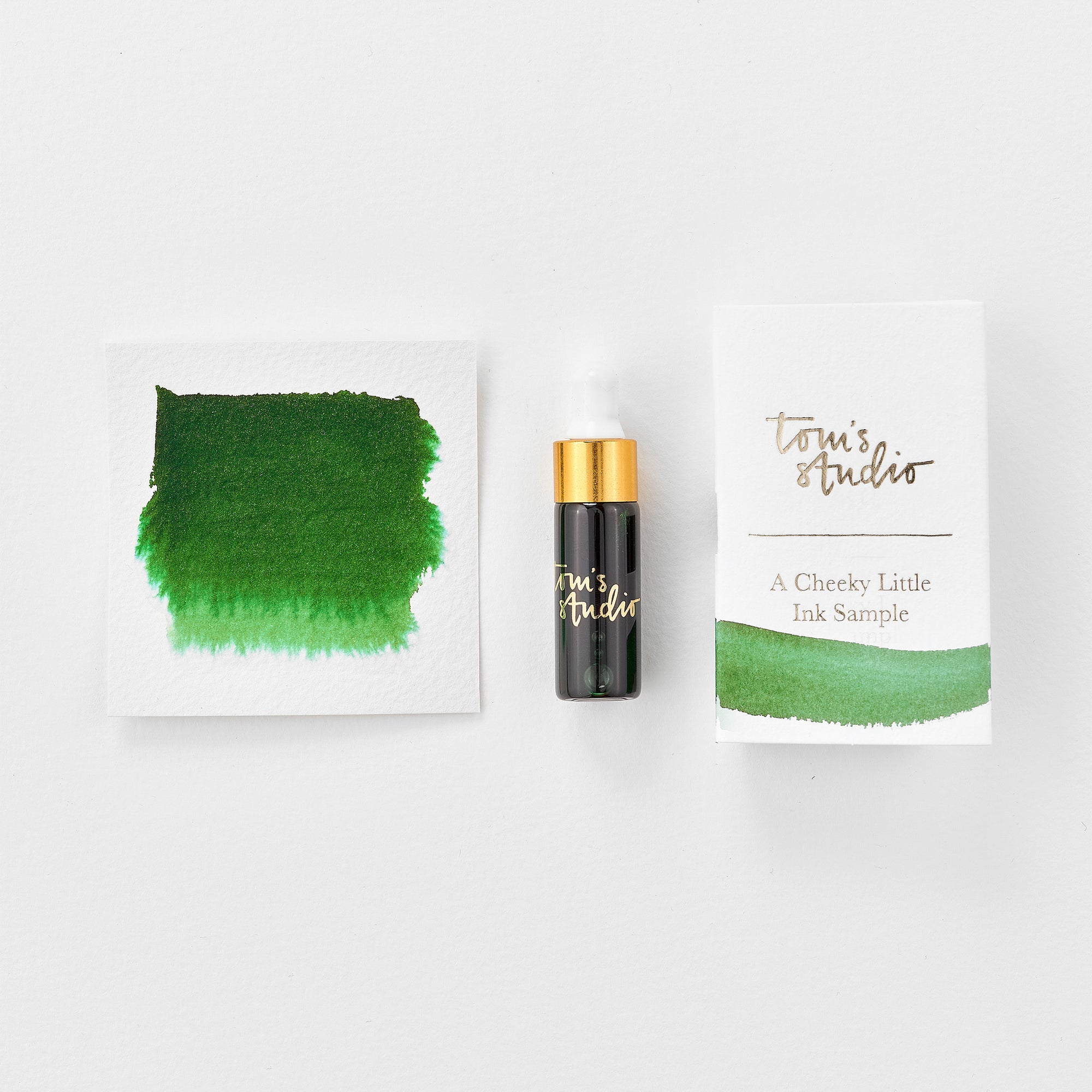 A mini ink sample bottle of New Forest Green fountain pen ink by Tom’s Studio, displayed alongside a swatch of the ink on textured paper and its matching packaging. The bottle is small, with a gold cap and a white pipette dropper, and features 'Tom’s Studio' in gold lettering. The ink swatch shows a rich, shimmering hue with a gradient from deep to lighter tones. The packaging, a small white card, has 'Tom’s Studio' embossed in gold at the top, with a brushstroke of the ink colour at the bottom.