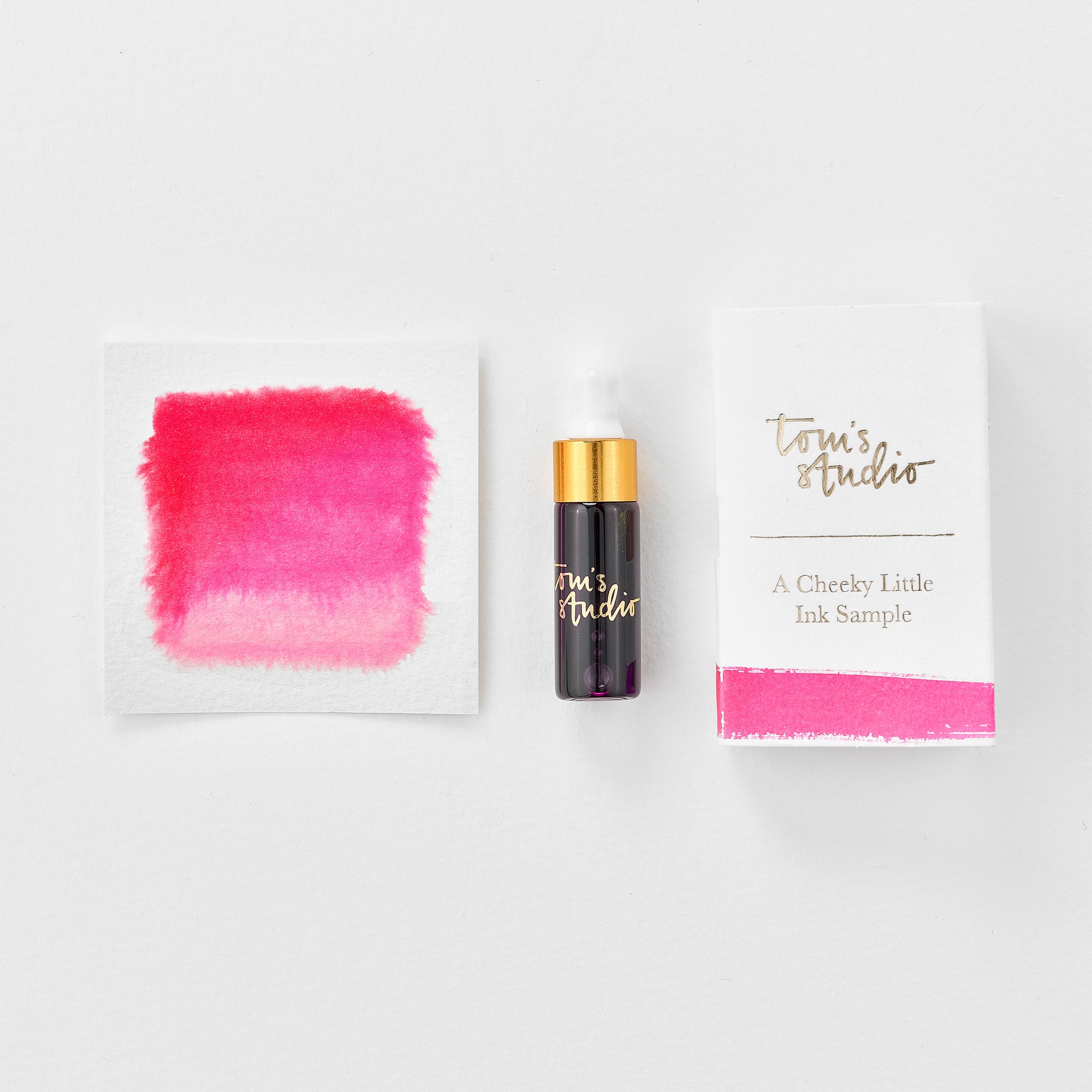 A mini ink sample bottle of Rambling Rose pink fountain pen ink by Tom’s Studio, displayed alongside a swatch of the ink on textured paper and its matching packaging. The bottle is small, with a gold cap and a white pipette dropper, and features 'Tom’s Studio' in gold lettering. The ink swatch shows a rich, shimmering hue with a gradient from deep to lighter tones. The packaging, a small white card, has 'Tom’s Studio' embossed in gold at the top, with a brushstroke of the ink colour at the bottom.