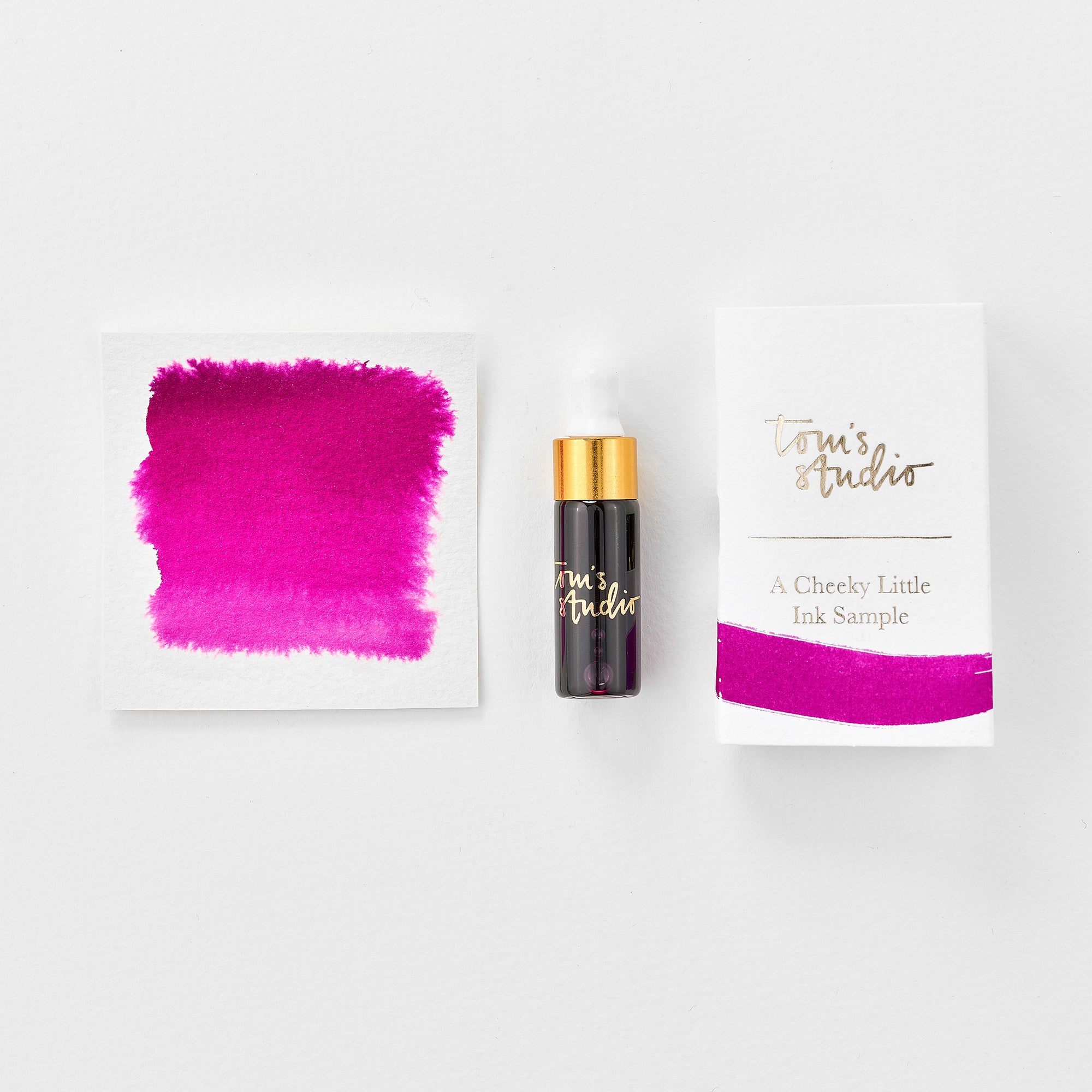 A mini ink sample bottle of Raspberry Sorbet pink fountain pen ink by Tom’s Studio, displayed alongside a swatch of the ink on textured paper and its matching packaging. The bottle is small, with a gold cap and a white pipette dropper, and features 'Tom’s Studio' in gold lettering. The ink swatch shows a rich, shimmering hue with a gradient from deep to lighter tones. The packaging, a small white card, has 'Tom’s Studio' embossed in gold at the top, with a brushstroke of the ink colour at the bottom.