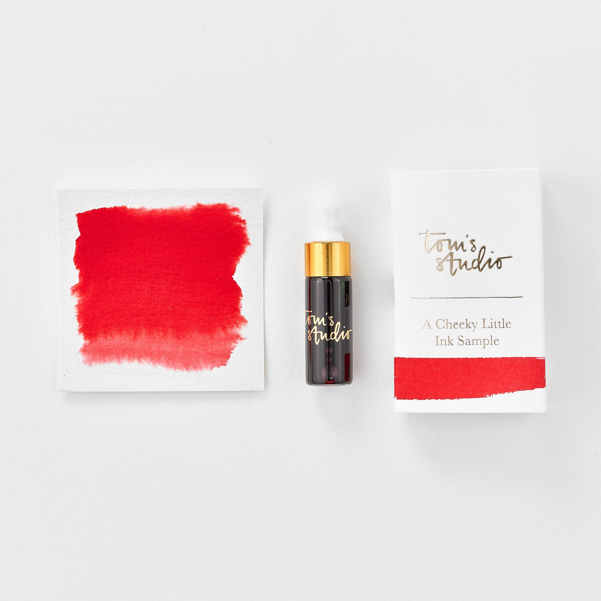 A mini ink sample bottle of Strawberry Jam red fountain pen ink by Tom’s Studio, displayed alongside a swatch of the ink on textured paper and its matching packaging. The bottle is small, with a gold cap and a white pipette dropper, and features 'Tom’s Studio' in gold lettering. The ink swatch shows a rich, shimmering hue with a gradient from deep to lighter tones. The packaging, a small white card, has 'Tom’s Studio' embossed in gold at the top, with a brushstroke of the ink colour at the bottom.