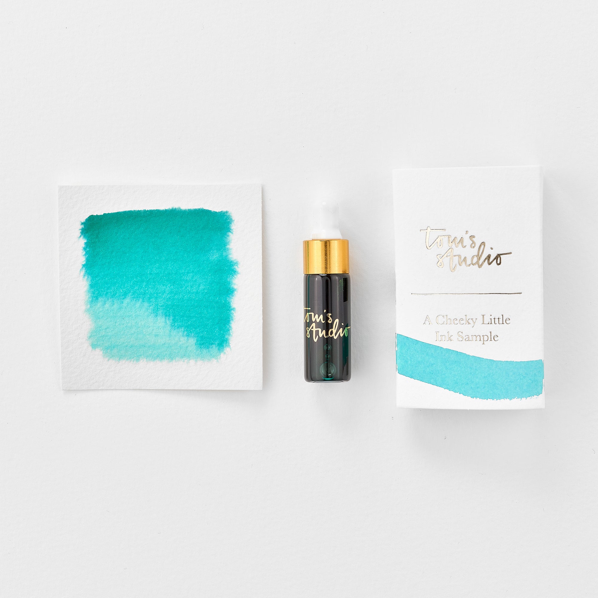 A mini ink sample bottle of Sunny Teal blue fountain pen ink by Tom’s Studio, displayed alongside a swatch of the ink on textured paper and its matching packaging. The bottle is small, with a gold cap and a white pipette dropper, and features 'Tom’s Studio' in gold lettering. The ink swatch shows a rich, shimmering hue with a gradient from deep to lighter tones. The packaging, a small white card, has 'Tom’s Studio' embossed in gold at the top, with a brushstroke of the ink colour at the bottom.