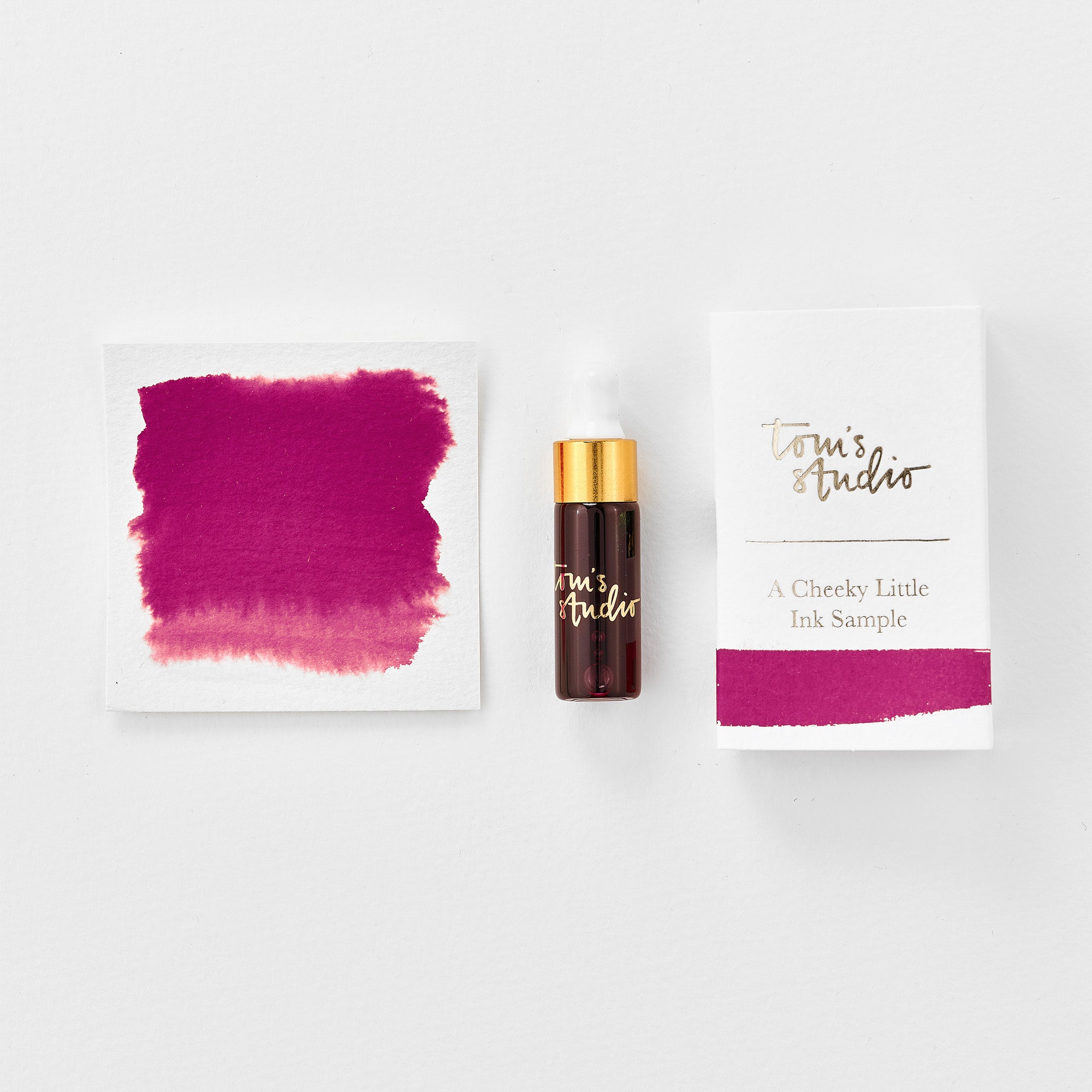 A mini ink sample bottle of Wild Cherry red/pink fountain pen ink by Tom’s Studio, displayed alongside a swatch of the ink on textured paper and its matching packaging. The bottle is small, with a gold cap and a white pipette dropper, and features 'Tom’s Studio' in gold lettering. The ink swatch shows a rich, shimmering hue with a gradient from deep to lighter tones. The packaging, a small white card, has 'Tom’s Studio' embossed in gold at the top, with a brushstroke of the ink colour at the bottom.