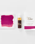 A mini ink sample bottle of Wild Cherry red/pink fountain pen ink by Tom’s Studio, displayed alongside a swatch of the ink on textured paper and its matching packaging. The bottle is small, with a gold cap and a white pipette dropper, and features 'Tom’s Studio' in gold lettering. The ink swatch shows a rich, shimmering hue with a gradient from deep to lighter tones. The packaging, a small white card, has 'Tom’s Studio' embossed in gold at the top, with a brushstroke of the ink colour at the bottom.