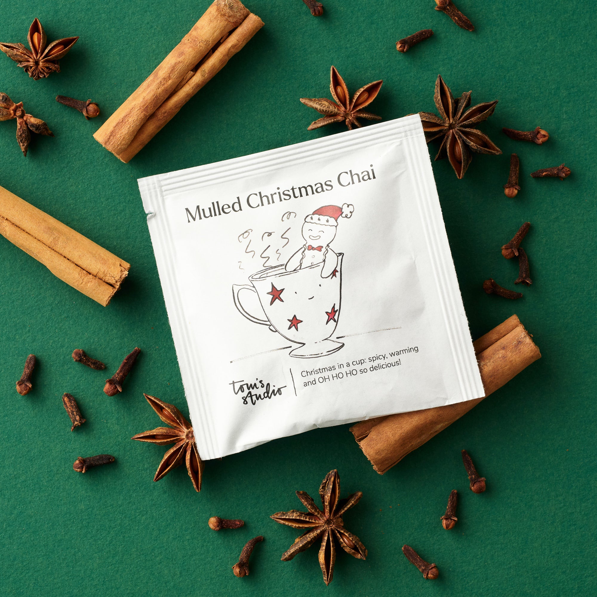 A sachet of Tom’s Studio Mulled Christmas Chai on a green background surrounded by cinnamon sticks, star anise, and cloves. The packet illustration shows a gingerbread figure in a teacup wearing a Santa hat, with the tagline “Christmas in a cup: spicy, warming and OH HO HO so delicious!”