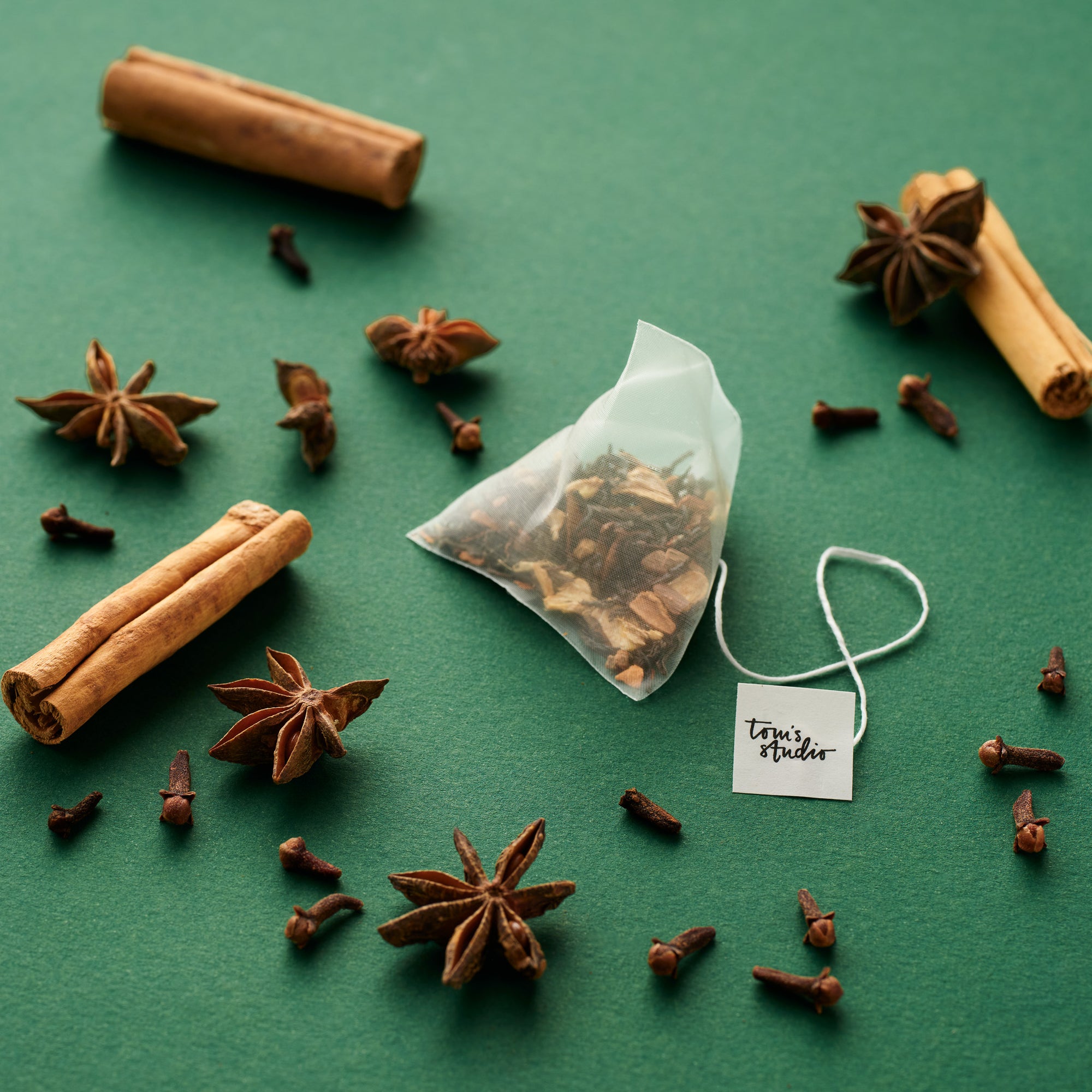 A Tom’s Studio Mulled Christmas Chai pyramid tea bag placed on a green background with scattered cinnamon sticks, star anise, and cloves. The tea tag features the Tom’s Studio logo.