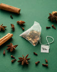 A Tom’s Studio Mulled Christmas Chai pyramid tea bag placed on a green background with scattered cinnamon sticks, star anise, and cloves. The tea tag features the Tom’s Studio logo.