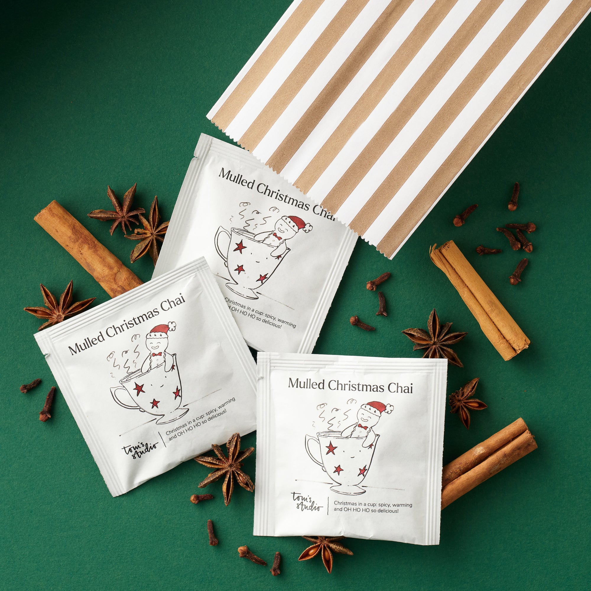 Three sachets of Tom’s Studio Mulled Christmas Chai arranged beside a striped paper bag on a green background, surrounded by festive spices including cinnamon sticks, star anise, and cloves.