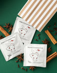 Three sachets of Tom’s Studio Mulled Christmas Chai arranged beside a striped paper bag on a green background, surrounded by festive spices including cinnamon sticks, star anise, and cloves.