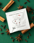 A sachet of Tom’s Studio Mulled Christmas Chai on a green background surrounded by cinnamon sticks, star anise, and cloves. The packet illustration shows a gingerbread figure in a teacup wearing a Santa hat, with the tagline “Christmas in a cup: spicy, warming and OH HO HO so delicious!”