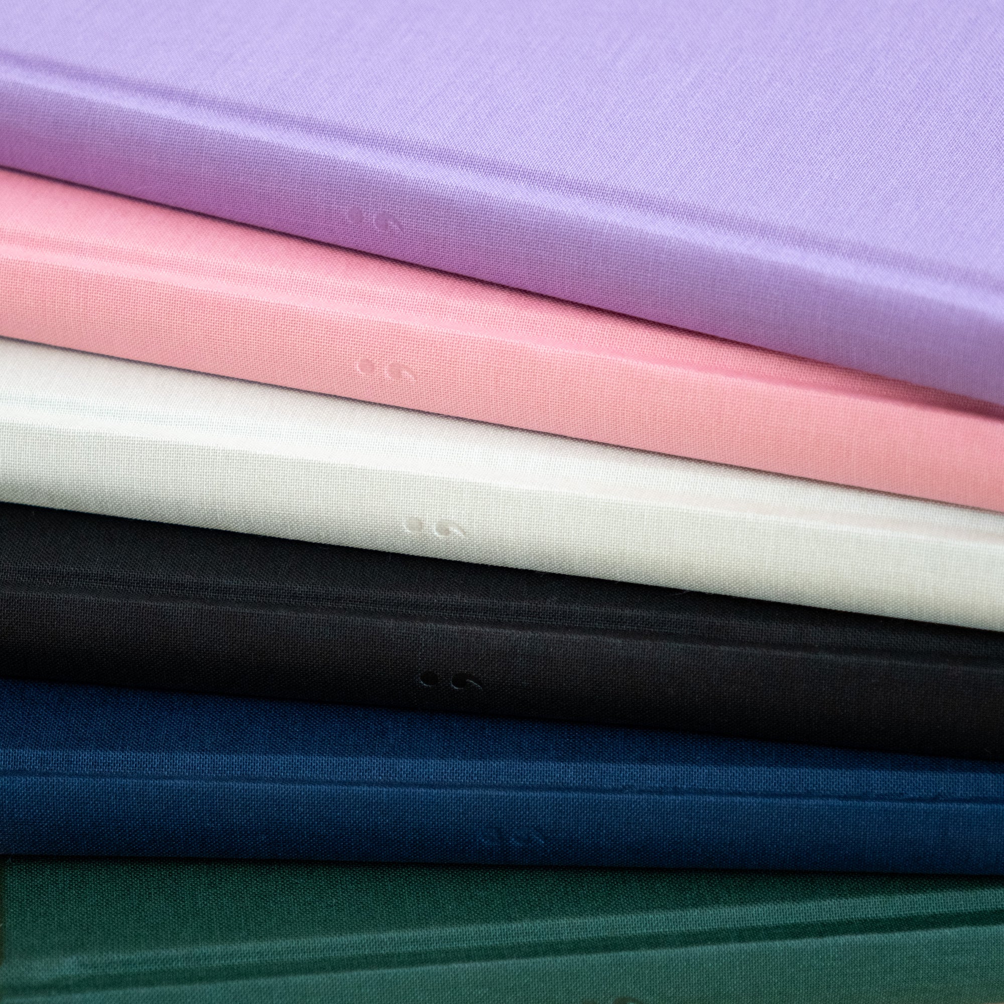 Detail of the embossed spine logo on a stack of vibrant Semikolon A5 notebooks in various colours, including Lilac, Flamingo pink, Moss green, Black, Marine blue and Forest green, with premium linen bound covers.