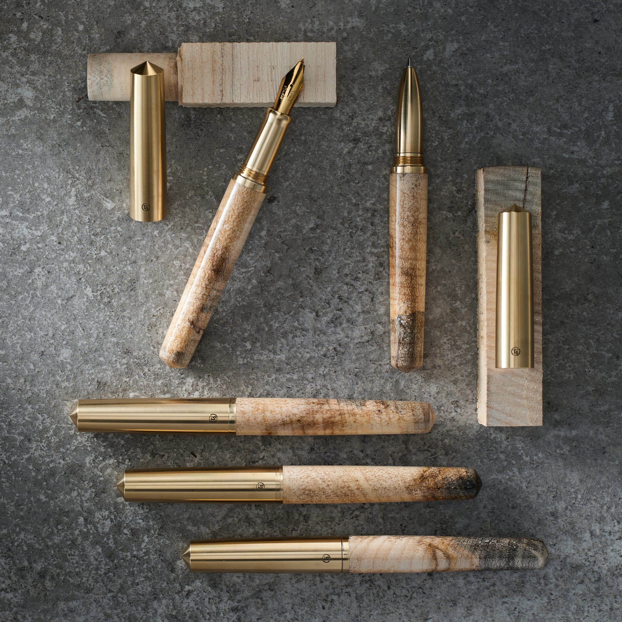 Four handmade sycamore fountain pens with marbled sycamore barrels and polished brass metal accents, arranged on a dark textured surface with matching gold pen stands.