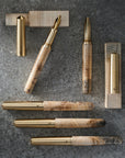 Four handmade sycamore fountain pens with marbled sycamore barrels and polished brass metal accents, arranged on a dark textured surface with matching gold pen stands.