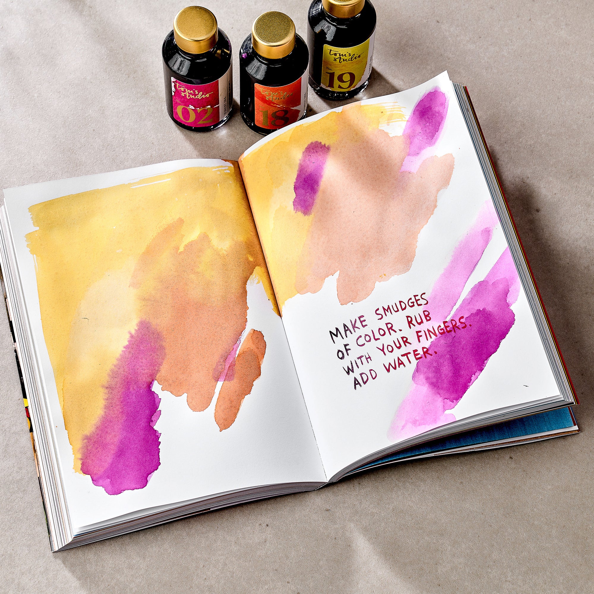 An open spread from Wreck This Journal, featuring colourful smudges of yellow, orange, pink, and purple ink on both pages. The right page includes the handwritten instruction: “MAKE SMUDGES OF COLOR. RUB WITH YOUR FINGERS. ADD WATER.” Three bottles of ink with gold caps are placed above the journal, each labelled with different numbers and colours.