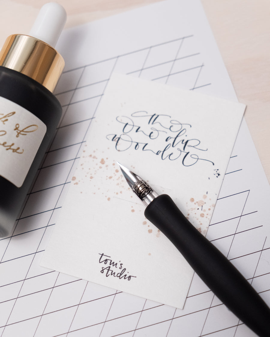 A straight nib calligraphy pen shown with writing on a notepad and spare ink