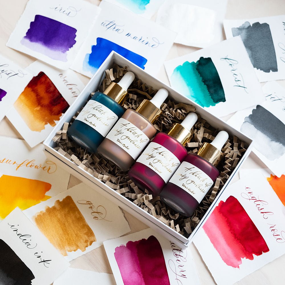 Four bottles of Tom's Studio Calligraphy Ink packaged in a gift box surrounded by ink swatches showing all available colours