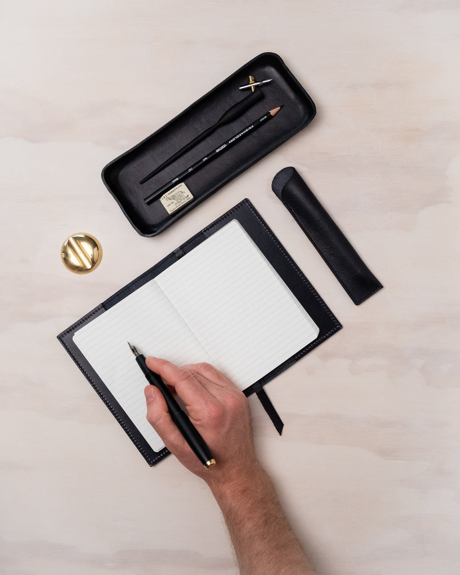 Writing in the black handmade leather journal with a fountain pen while a desk tray and pen rest store an oblique calligraphy pen